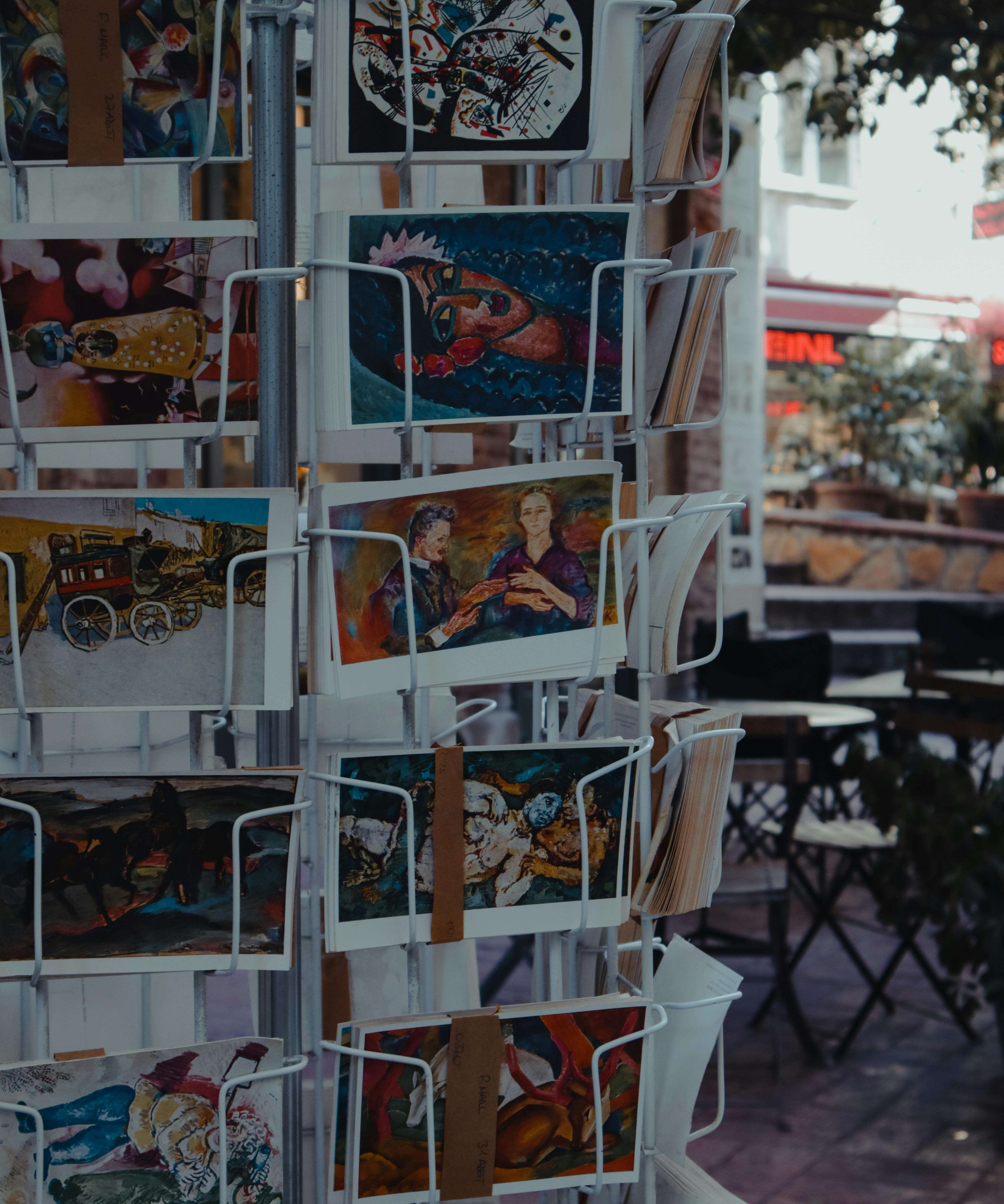A variety of colorful artistic postcards displayed on a metal rack in an outdoor setting.