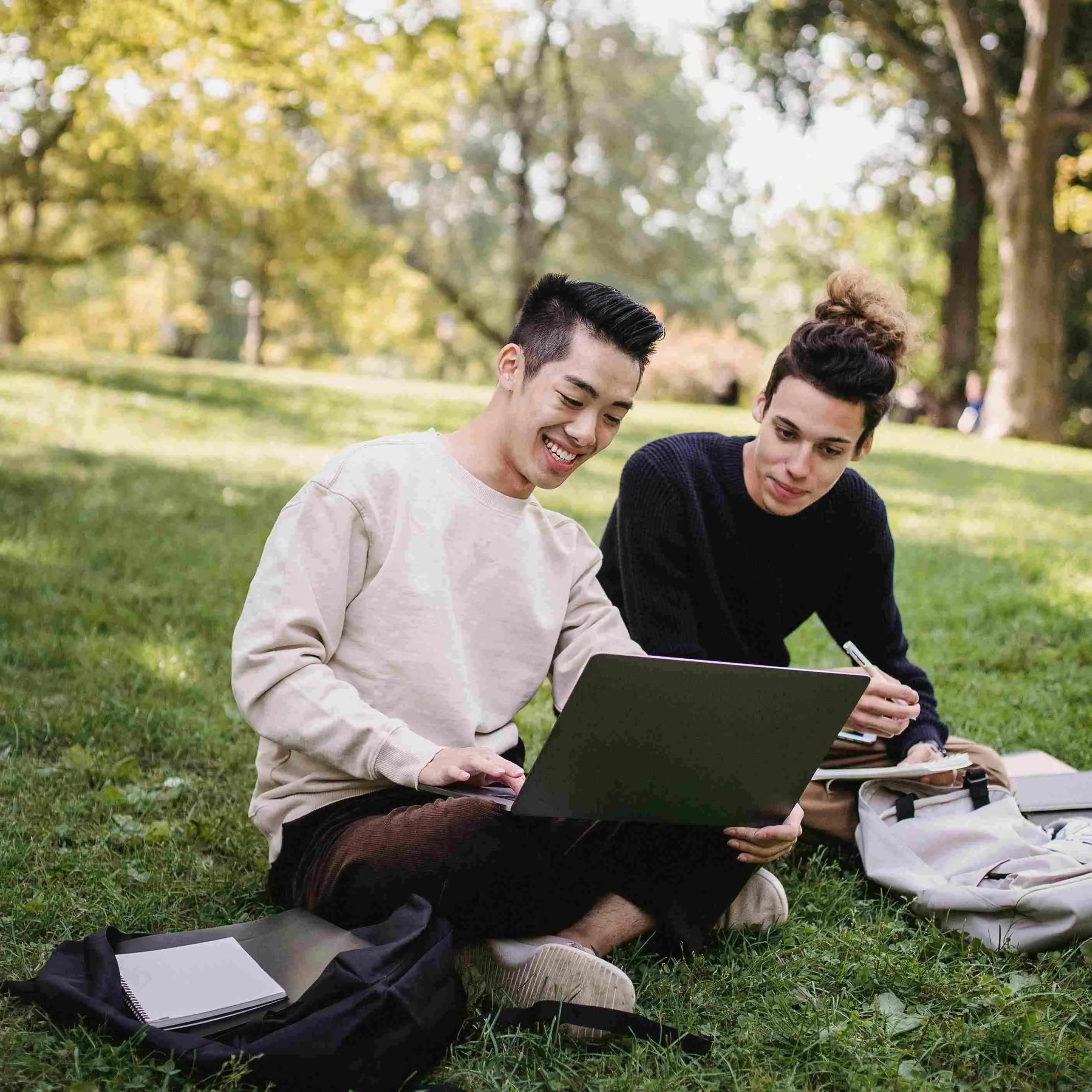 Full length content ethnic male students in casual outfits browsing laptop while sitting on meadow w